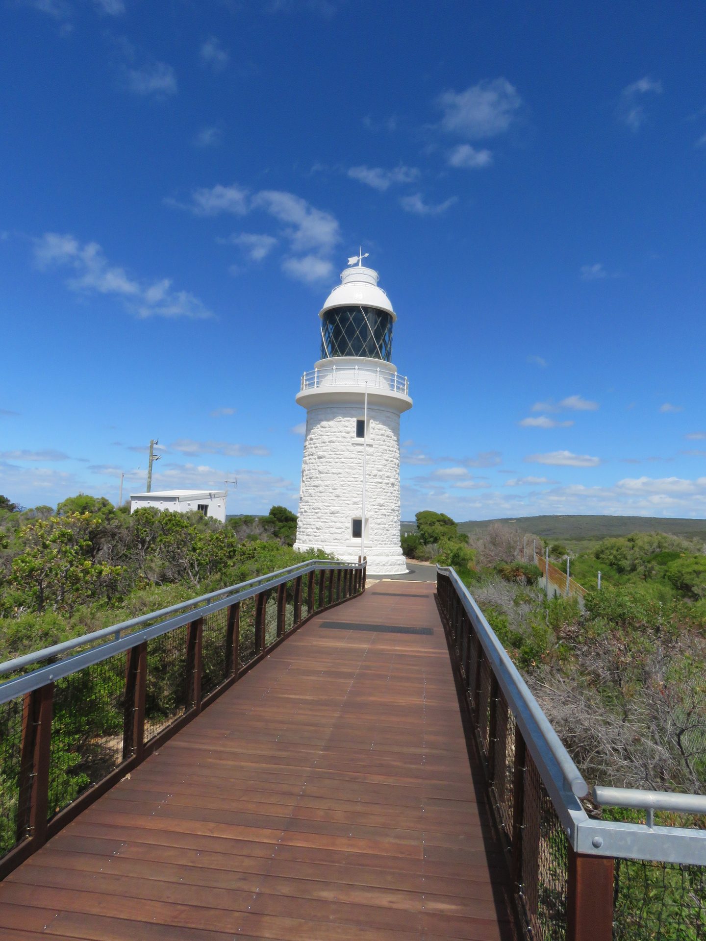 Ein Holzsteg führt durch niedrige Küstenvegetation zum weißen Leuchtturm am Cape Naturaliste.